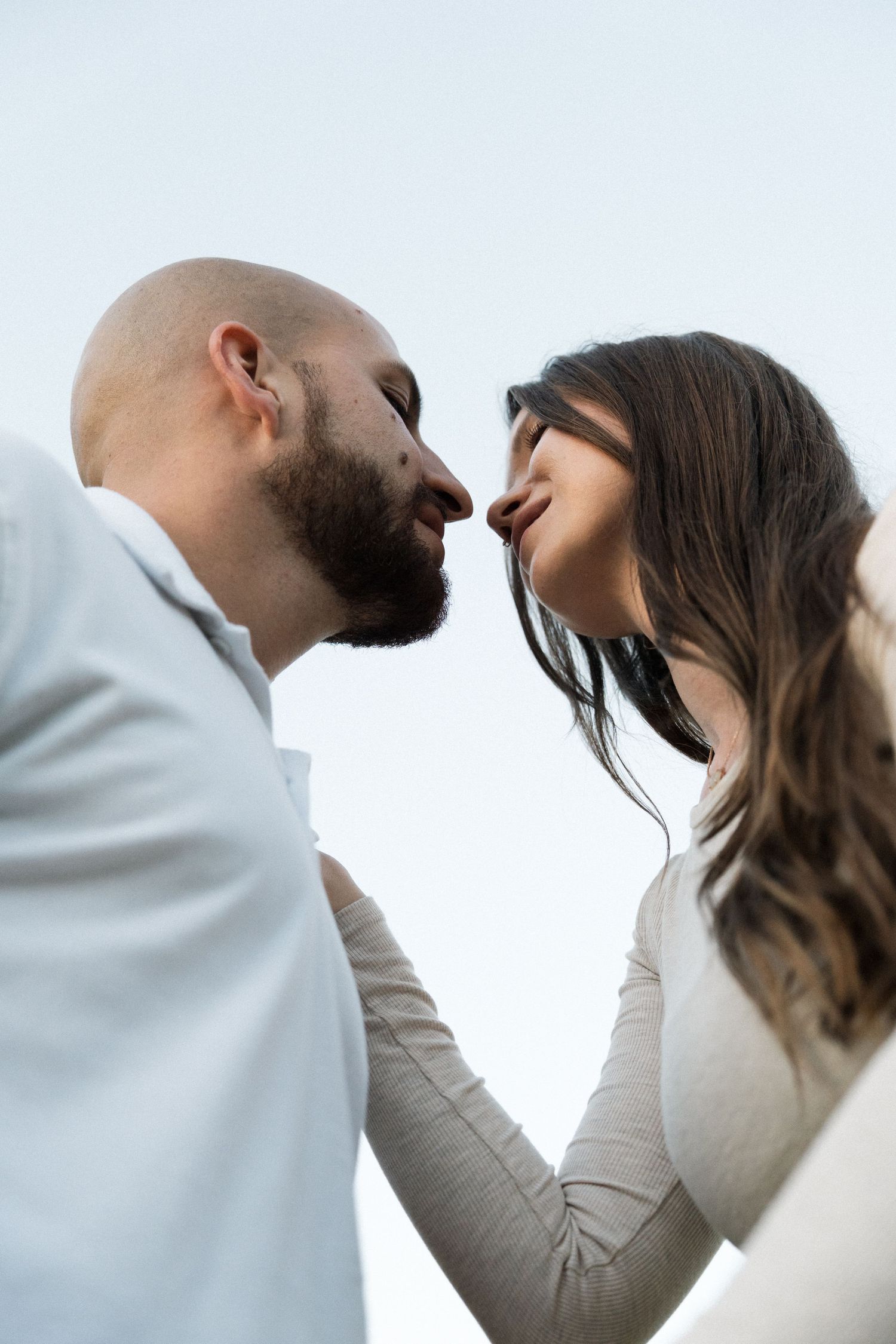 Low-angle close-up of a bearded man and a long-haired woman nearly touching noses in a romantic moment against a soft pale sky.