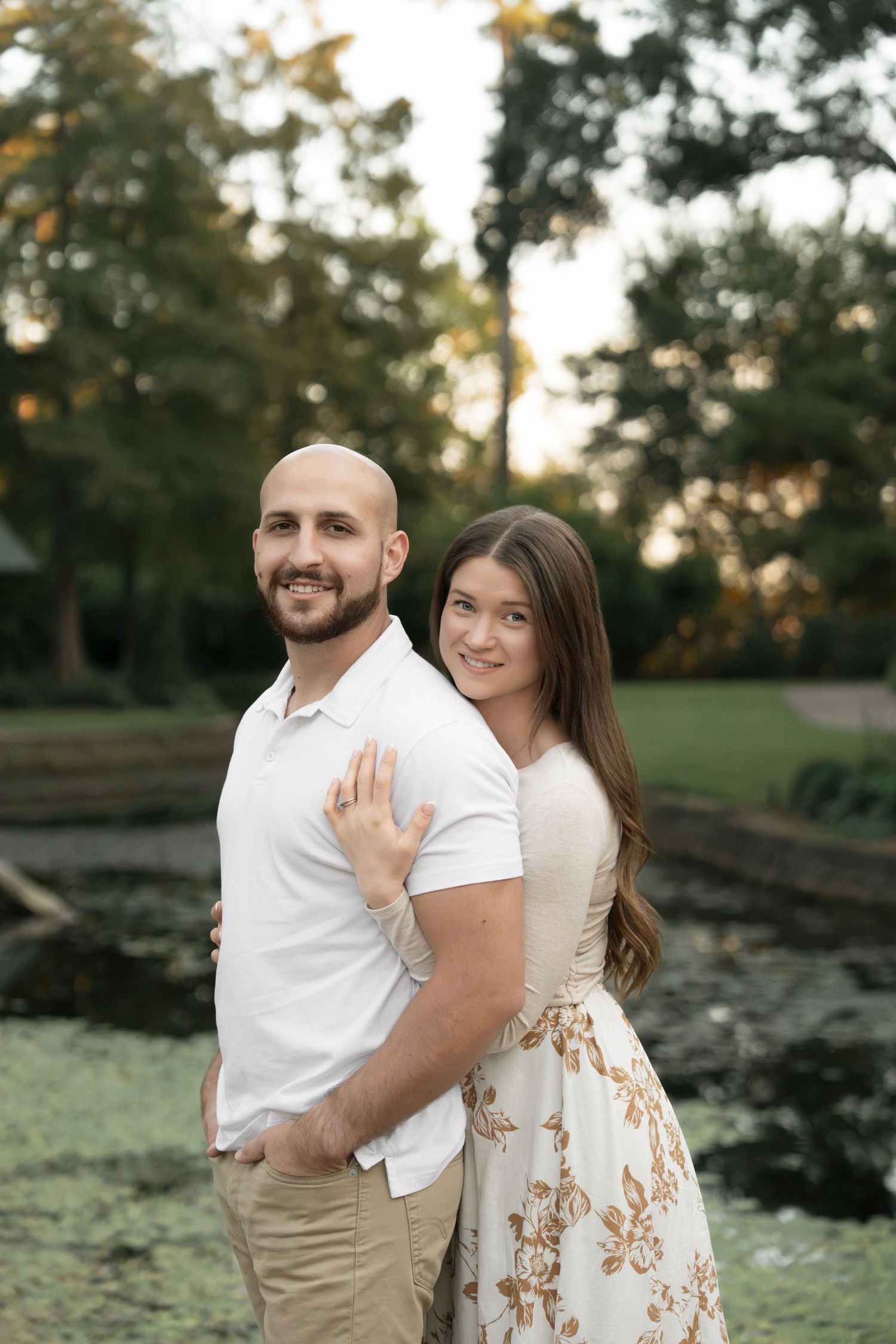 A smiling couple poses outdoors for an engagement photo, the woman hugging the man from behind beside a tranquil pond and trees.