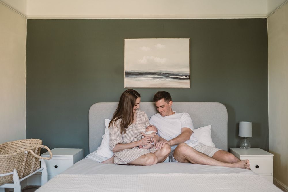 Young couple sitting on bed admiring their newborn baby in a modern bedroom with sage green walls.