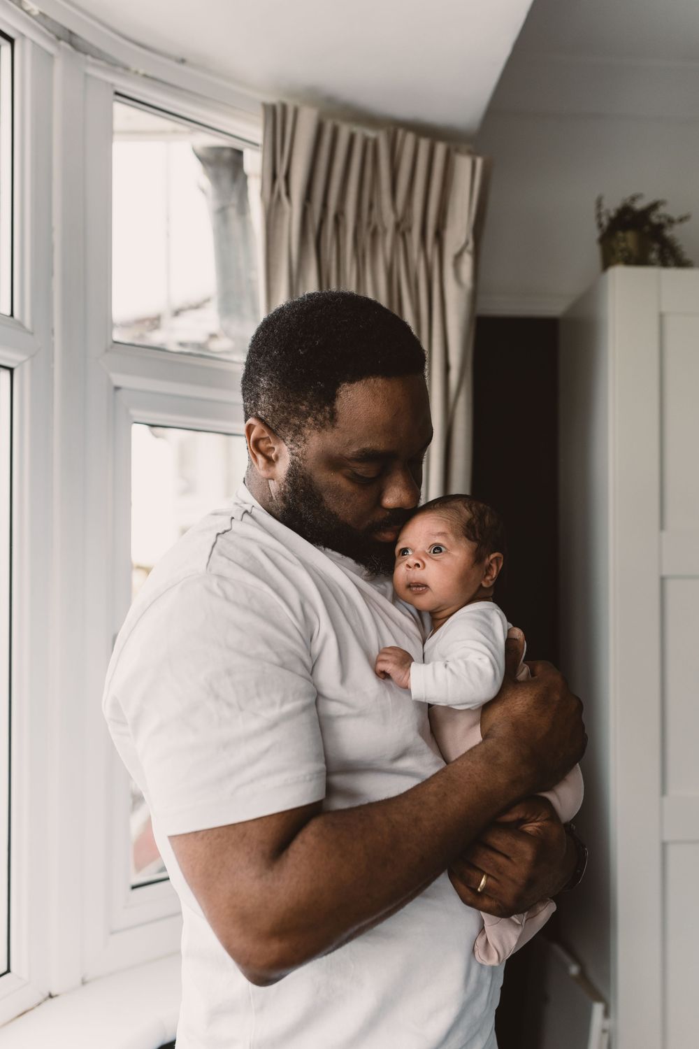 A father tenderly cuddles his newborn baby while standing by a bright window indoors.
