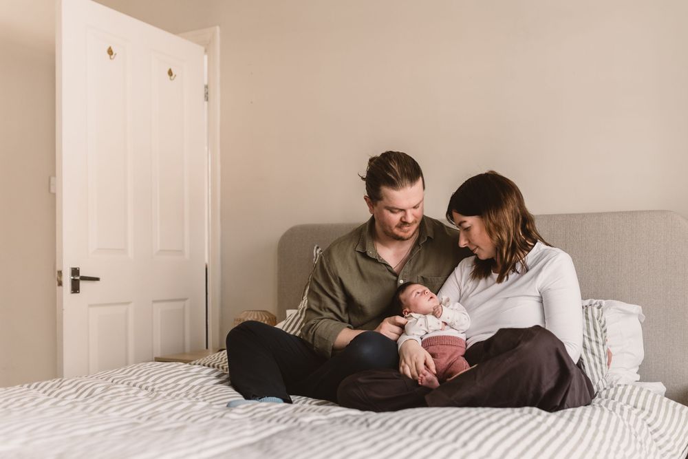 Happy couple sitting on bed together admiring their newborn baby in a bright, cozy bedroom.