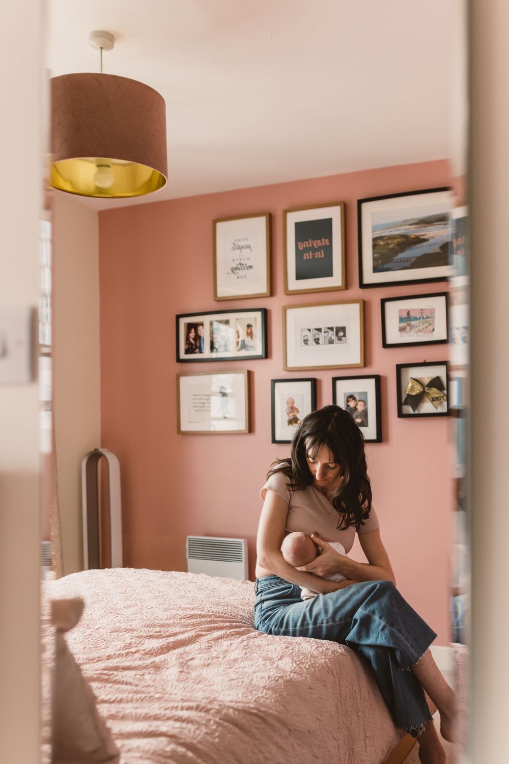 A mother sits on a pink bed cradling her newborn baby in a cozy bedroom with a gallery wall.