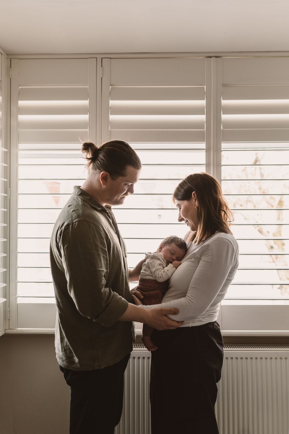 A couple stands by shuttered windows, lovingly gazing at their newborn baby held in the mother's arms.