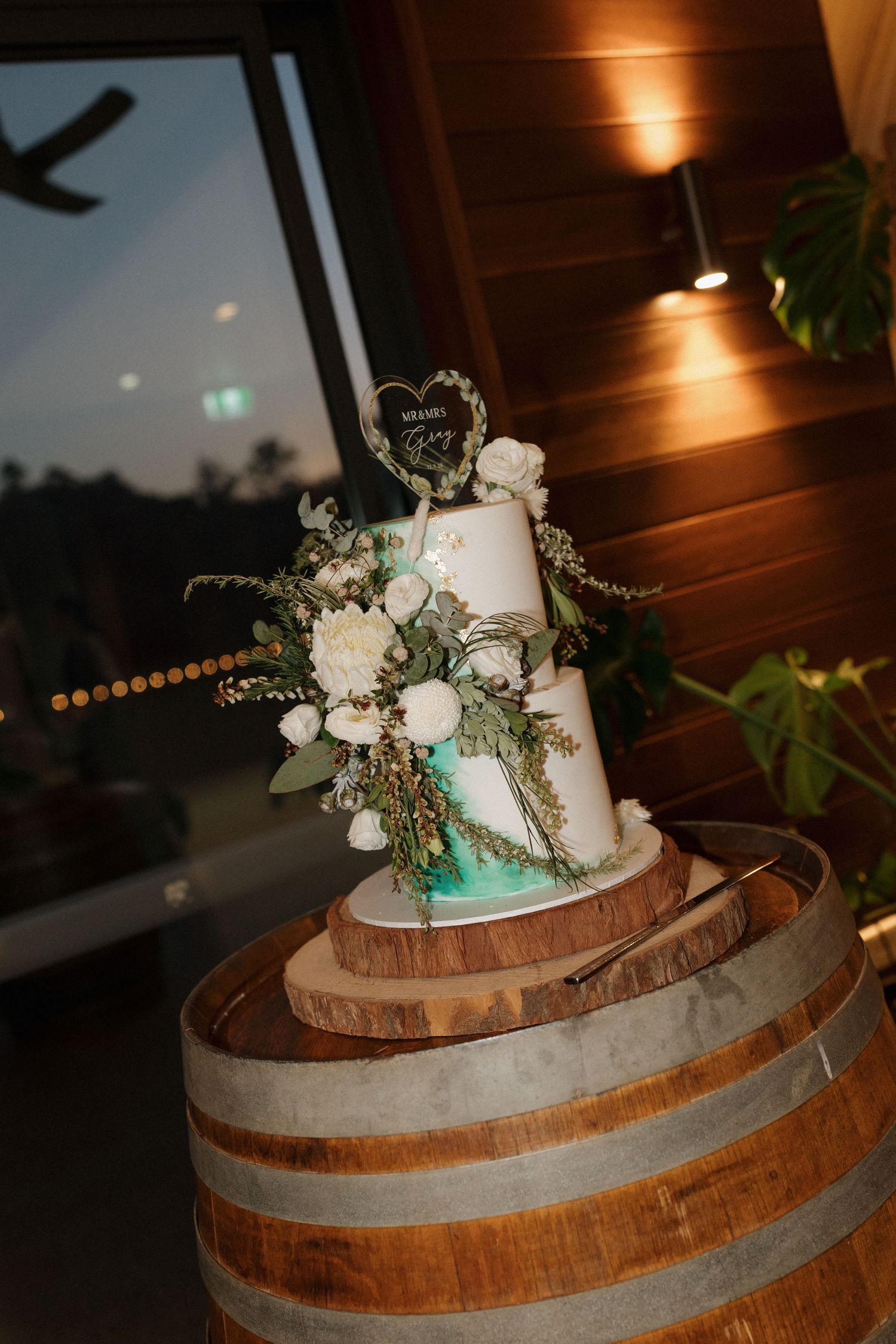 Two-tier white wedding cake with white florals and greenery on a rustic wooden barrel in warm evening venue lighting.