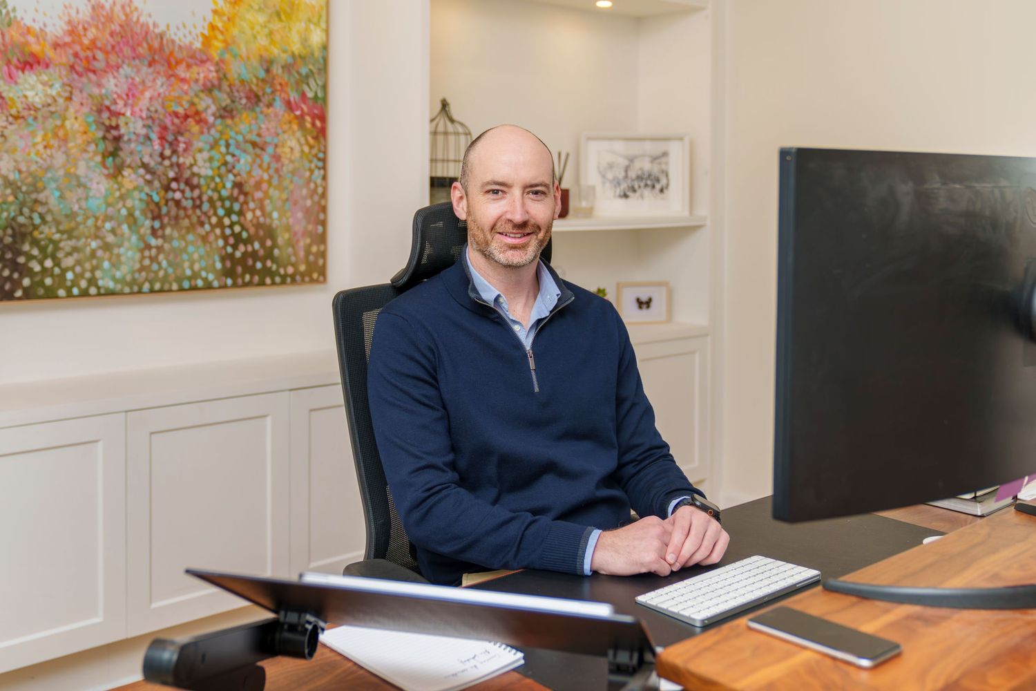 Bald man in navy sweater reading a book at a modern home office desk with large windows and greenery.