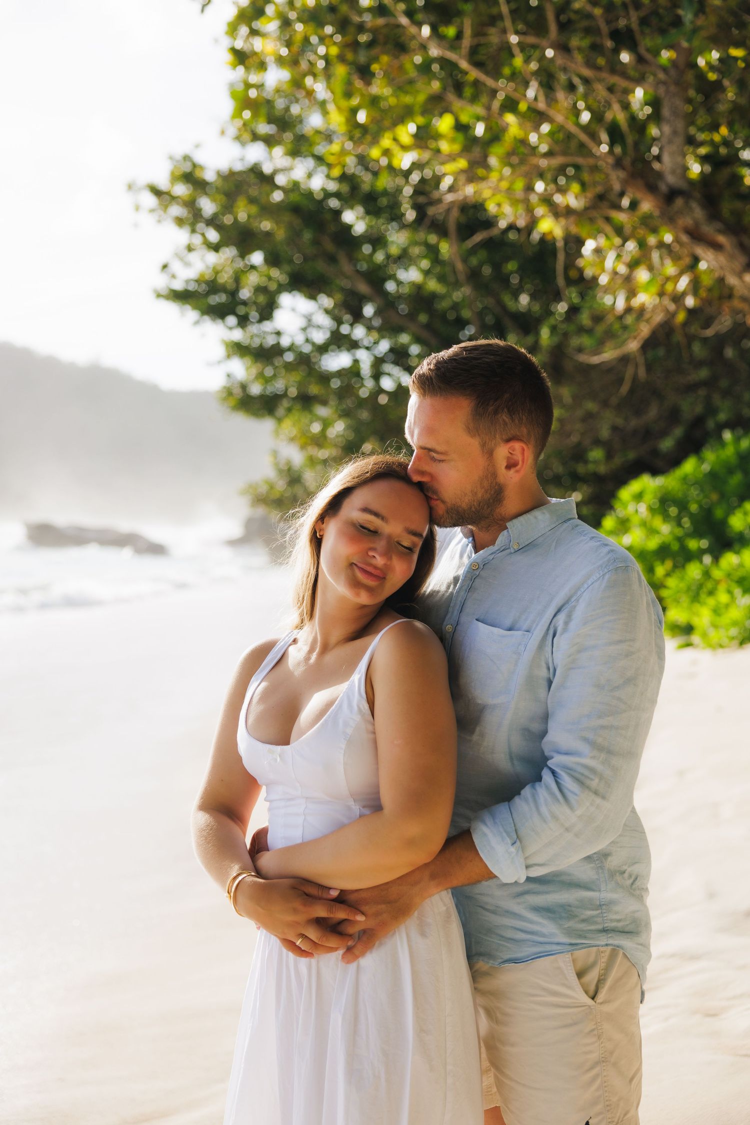 A couple shares a tender embrace on a tropical beach, the man kissing the womans head as she smiles.