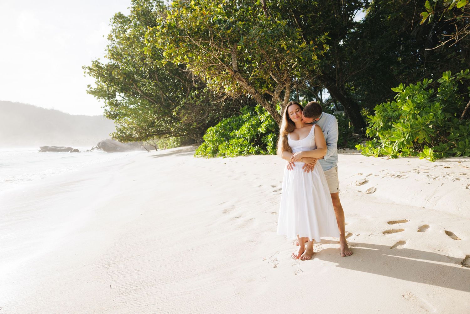 A couple in white outfits walk hand in hand on a pristine tropical beach surrounded by lush greenery.