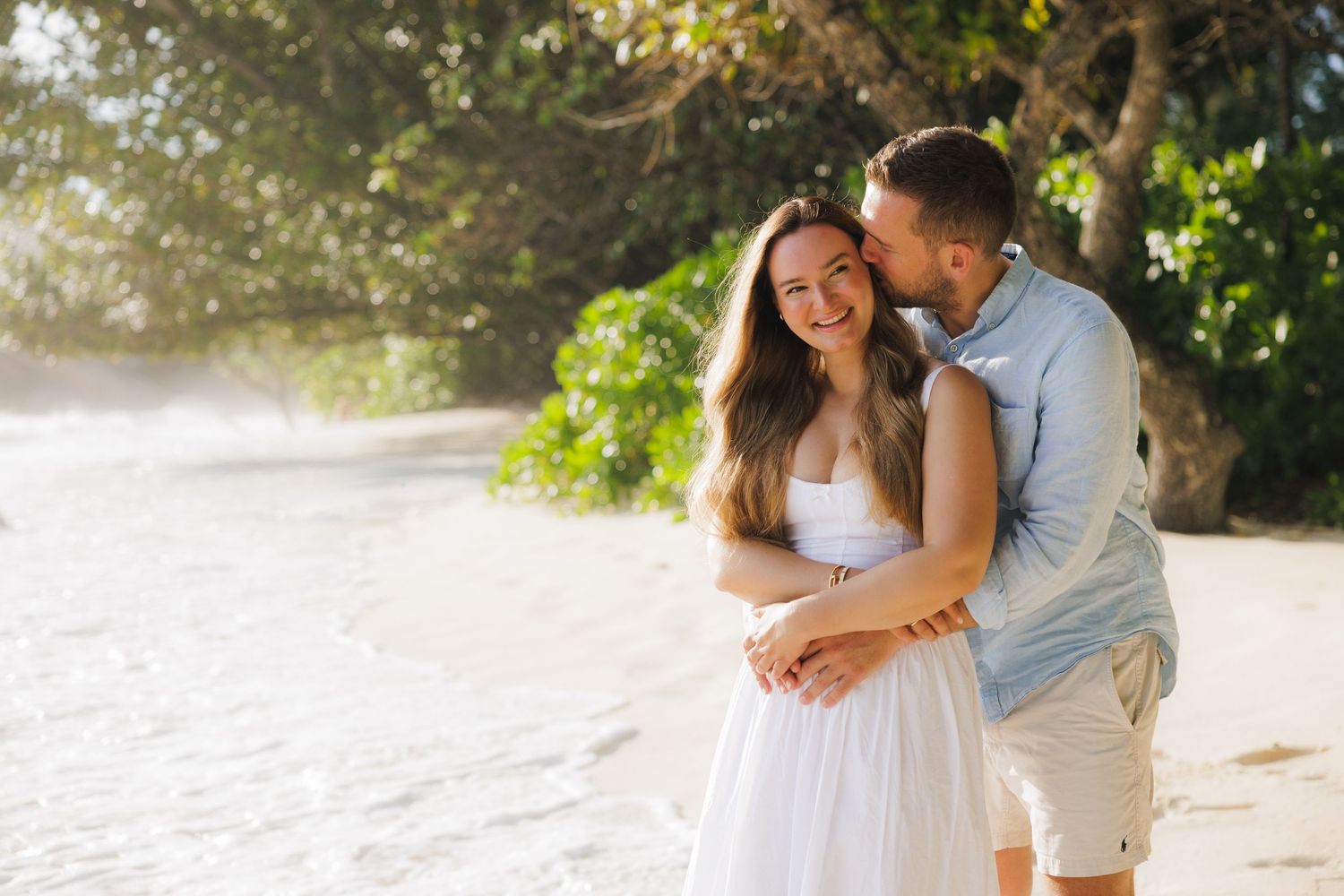 A smiling woman in white enjoys a romantic kiss on the cheek from her partner on a sunny tropical beach.