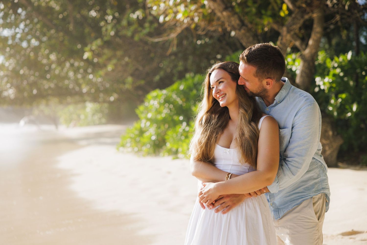 A happy couple laughs and embraces on a sunlit tropical beach, the man whispering in the womans ear.