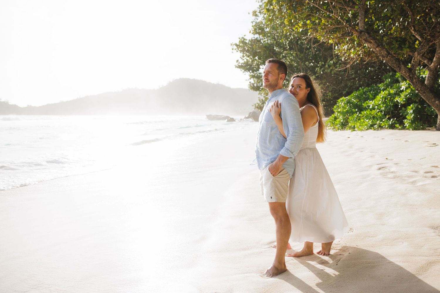 A couple shares a loving embrace on a bright sandy tropical beach with lush green trees behind them.