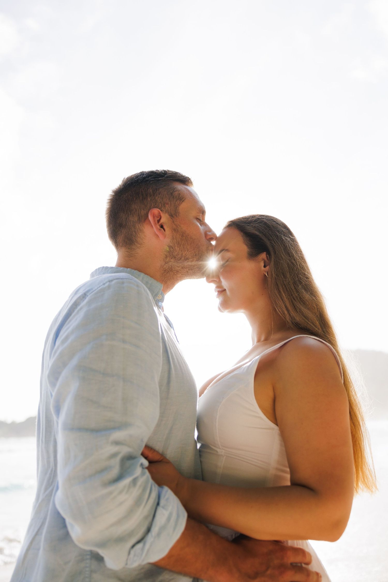 A romantic couple shares a tender kiss on a sun-drenched tropical beach with bright backlight glowing.