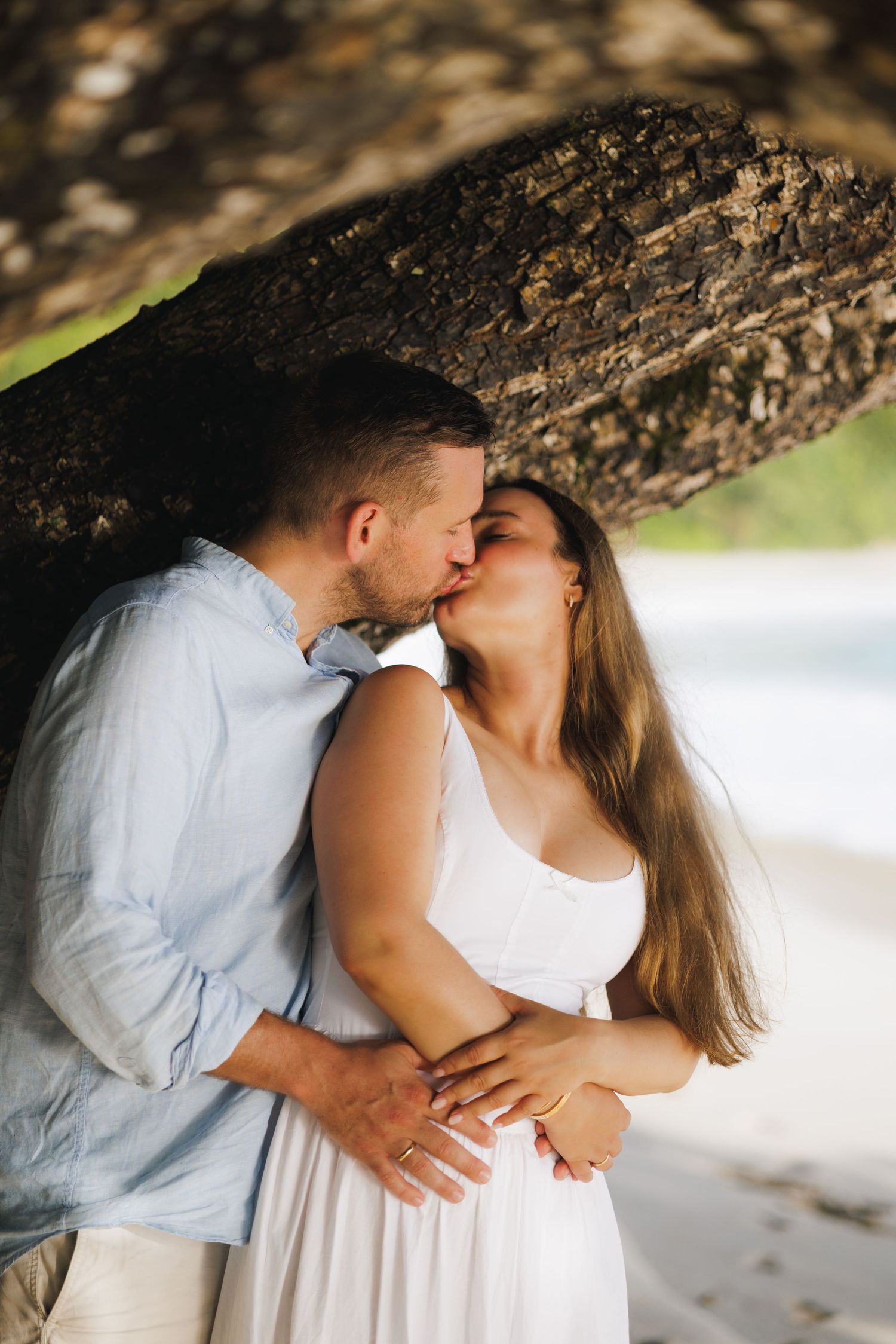 A couple shares a passionate kiss under a large tree trunk on a beautiful tropical sandy beach.