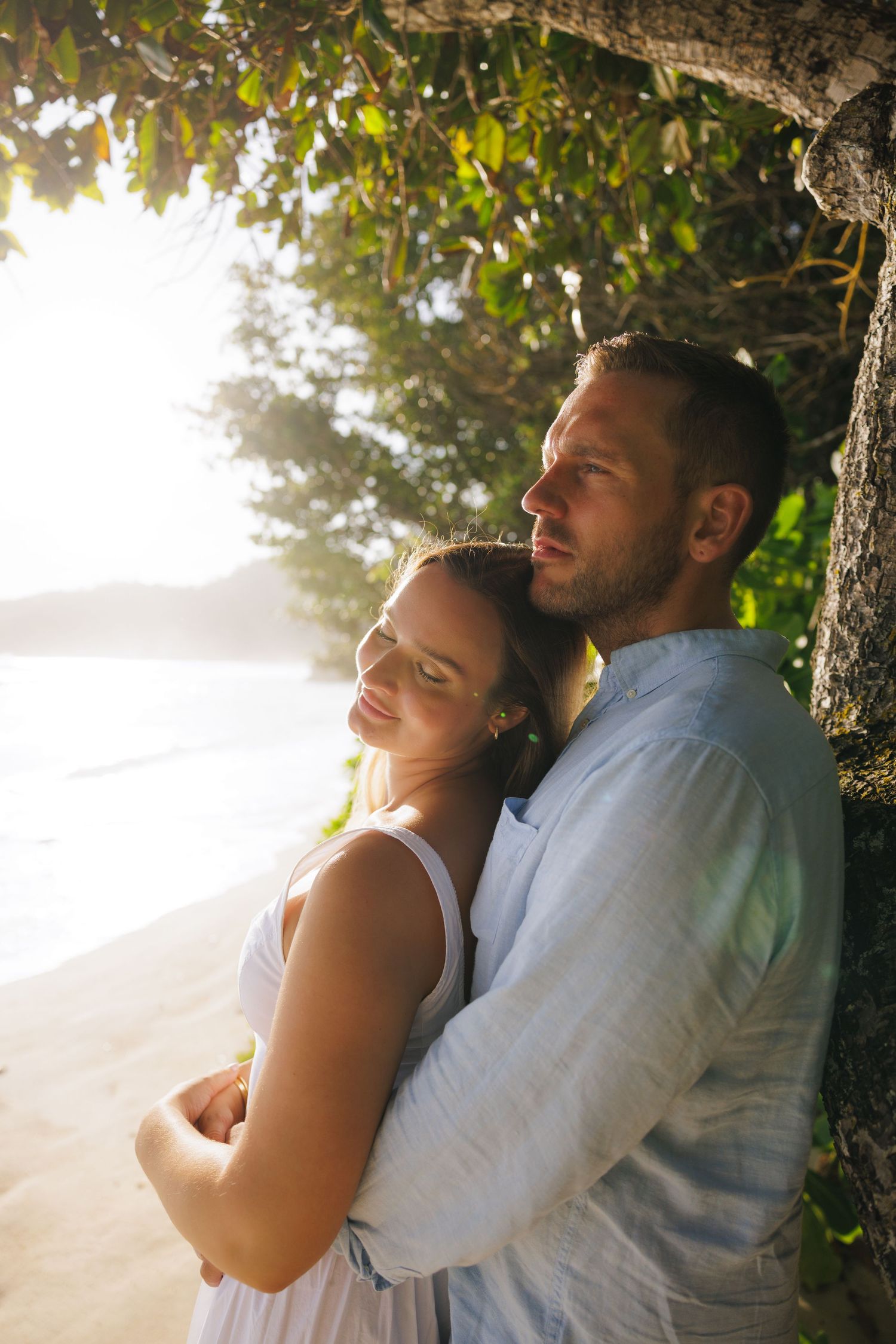 Couple embracing under a tree on a tropical beach at golden sunset, woman in white dress leaning on mans chest.