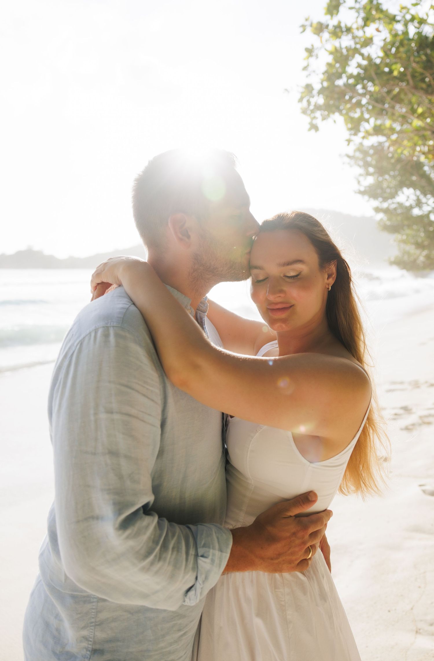 A couple embraces tenderly on a sunny tropical beach, the man kissing the womans cheek in warm sunlight.