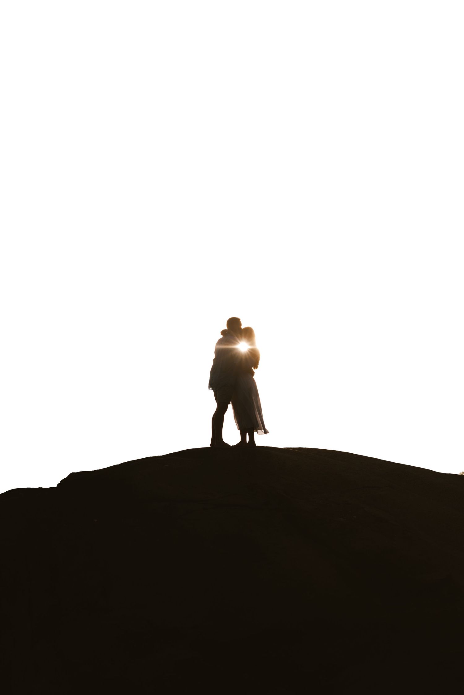 Romantic couple silhouette embracing on hilltop against bright white sky at sunset.