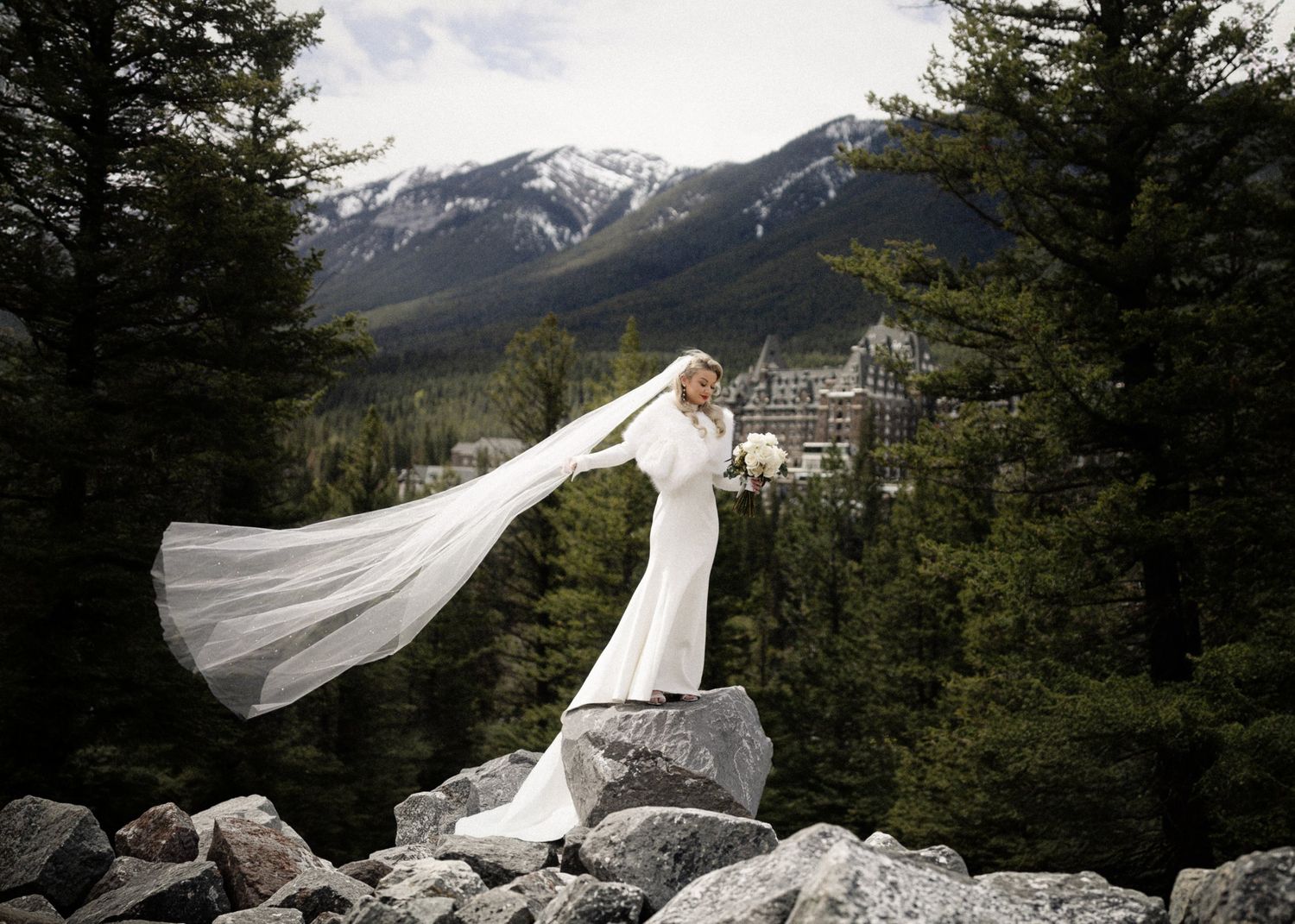 Bride in white gown with flowing veil on rocky mountain landscape, Banff Springs Hotel visible among evergreen trees.