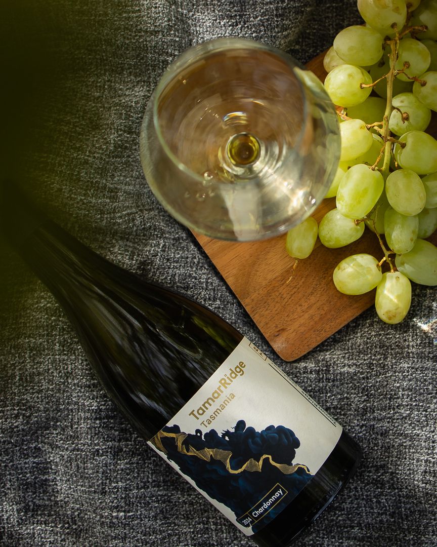 Tamar Ridge Tasmania Chardonnay wine bottle beside an empty glass and fresh green grapes on a wooden board.