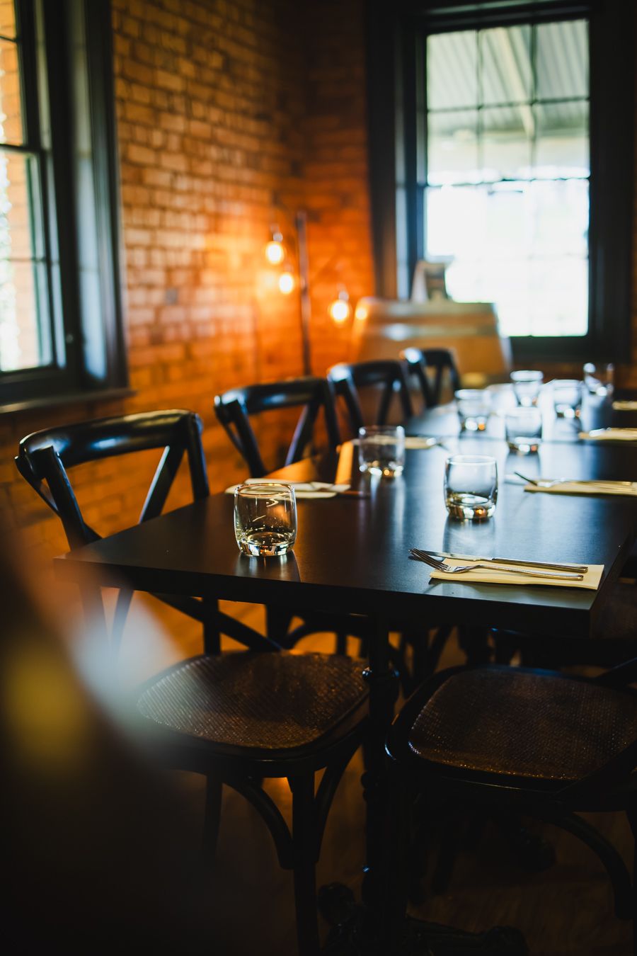 Elegant restaurant interior with dark wood tables, cross-back chairs, warm Edison bulb lighting, and brick walls.