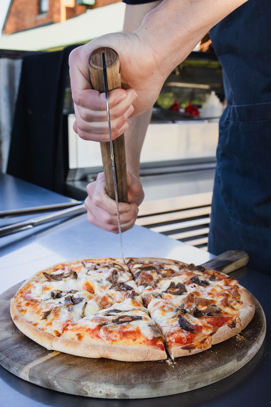 Chef slicing a freshly baked mushroom pizza on a wooden board using a traditional wooden-handled pizza cutter.
