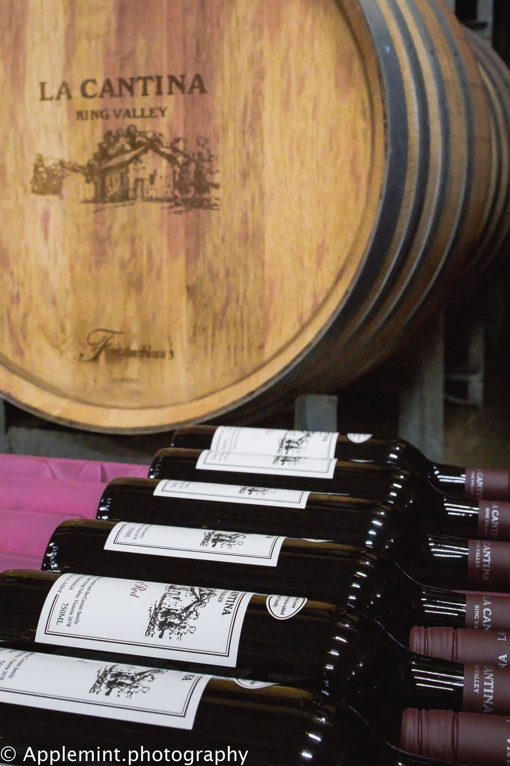 Wine bottles stacked in front of a La Cantina King Valley oak barrel in a winery cellar.