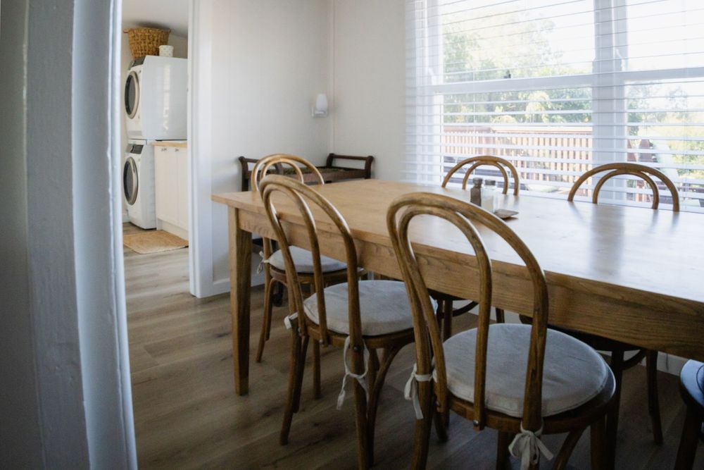 Rustic farmhouse dining room with wooden table, bentwood chairs, large window, and stacked washer-dryer visible in background.