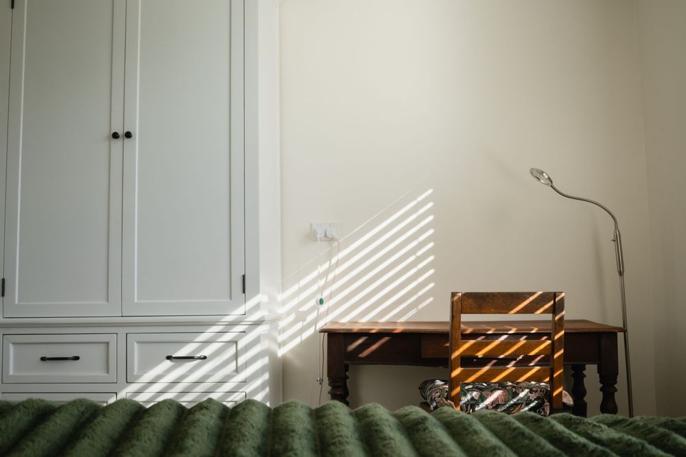 Sunlit bedroom corner with white wardrobe, wooden side table, floor lamp, and green textured bedspread.