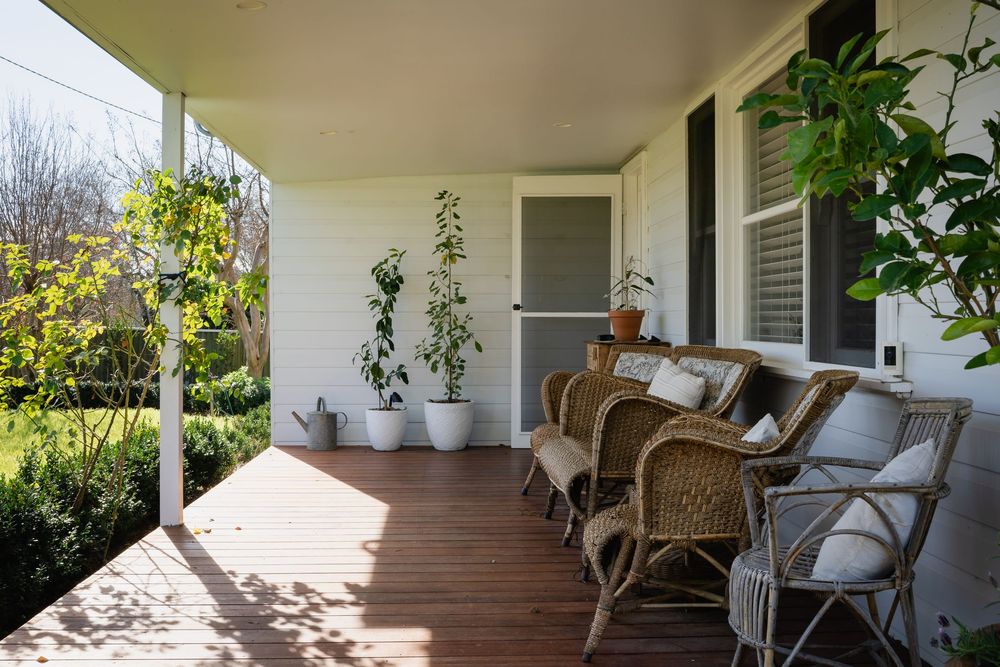 Charming farmhouse porch with wicker chairs, potted plants, and warm timber decking surrounded by lush greenery.