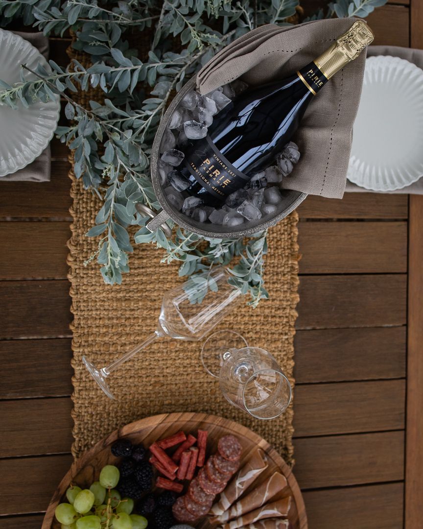 Overhead view of charcuterie board with grapes, champagne bottle on ice, and two glasses on a woven table runner.