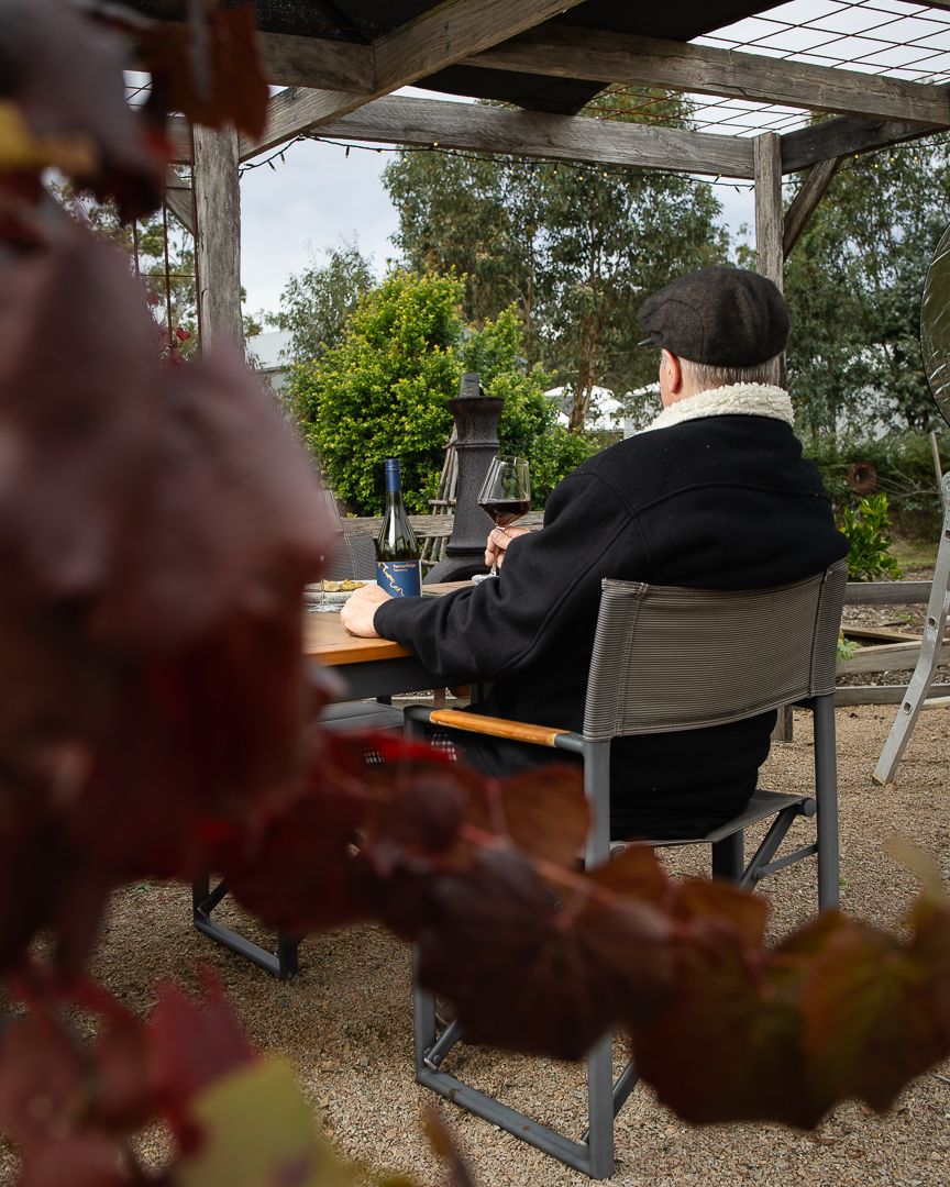 Person sitting outdoors under a pergola at a rustic table, surrounded by autumn foliage at a winery.
