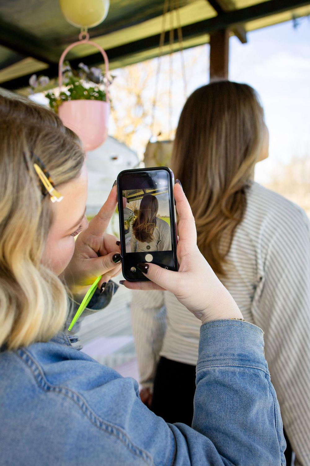 Woman in denim jacket photographs friend with smartphone at outdoor cafe with hanging plants.