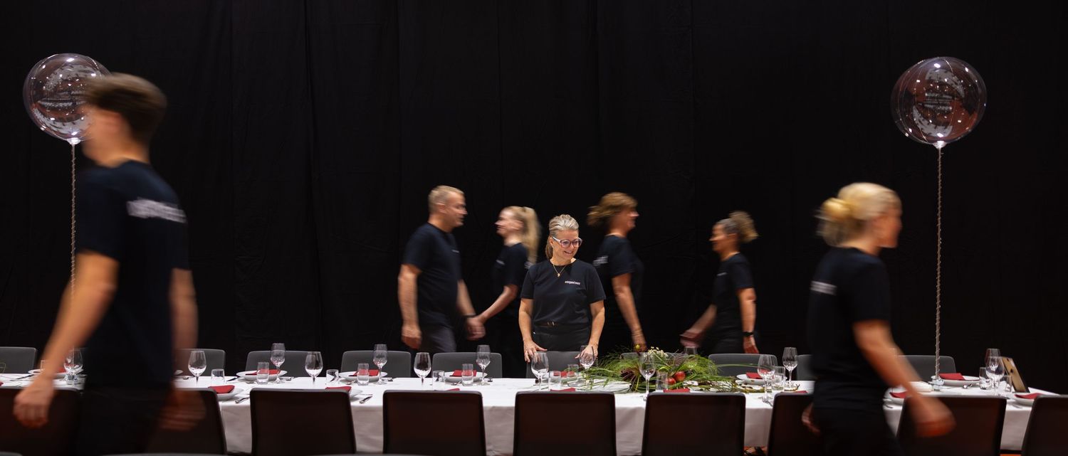 Staff in black uniforms stand behind an elegantly set banquet table with disco balls and floral centerpieces in a dark venue.