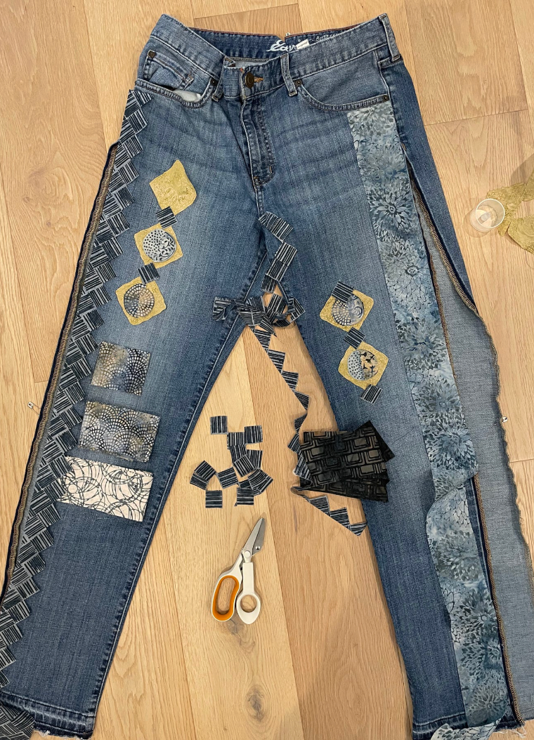 DIY upcycled jeans with patchwork fabric squares in gold and blue, displayed on wood floor with scissors nearby.