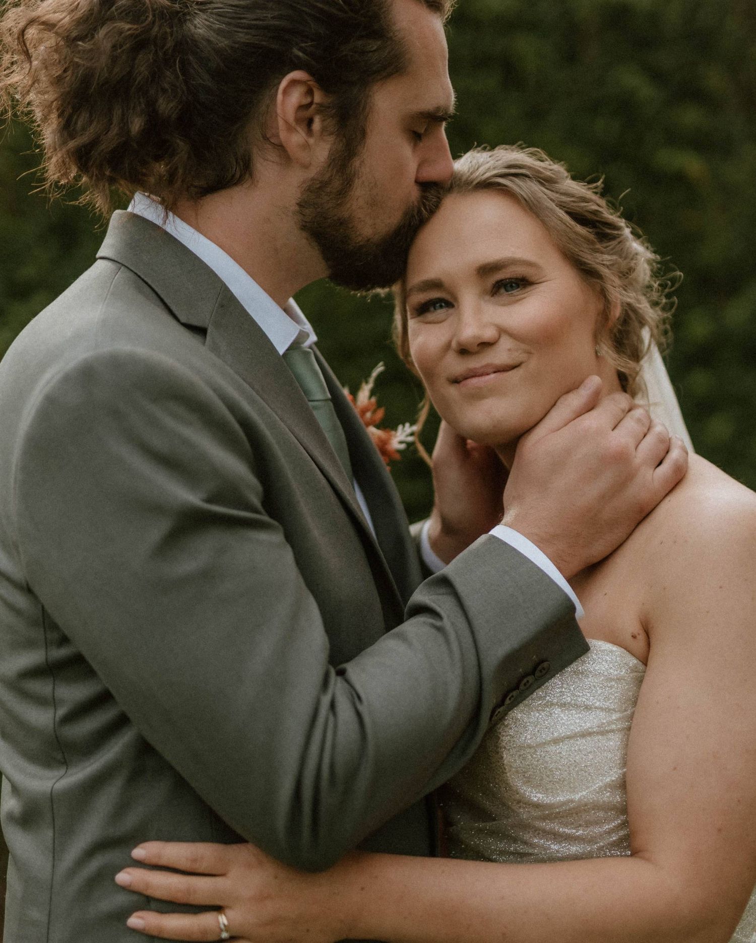 Groom in gray suit kisses bride on forehead while tenderly holding her face on their wedding day.