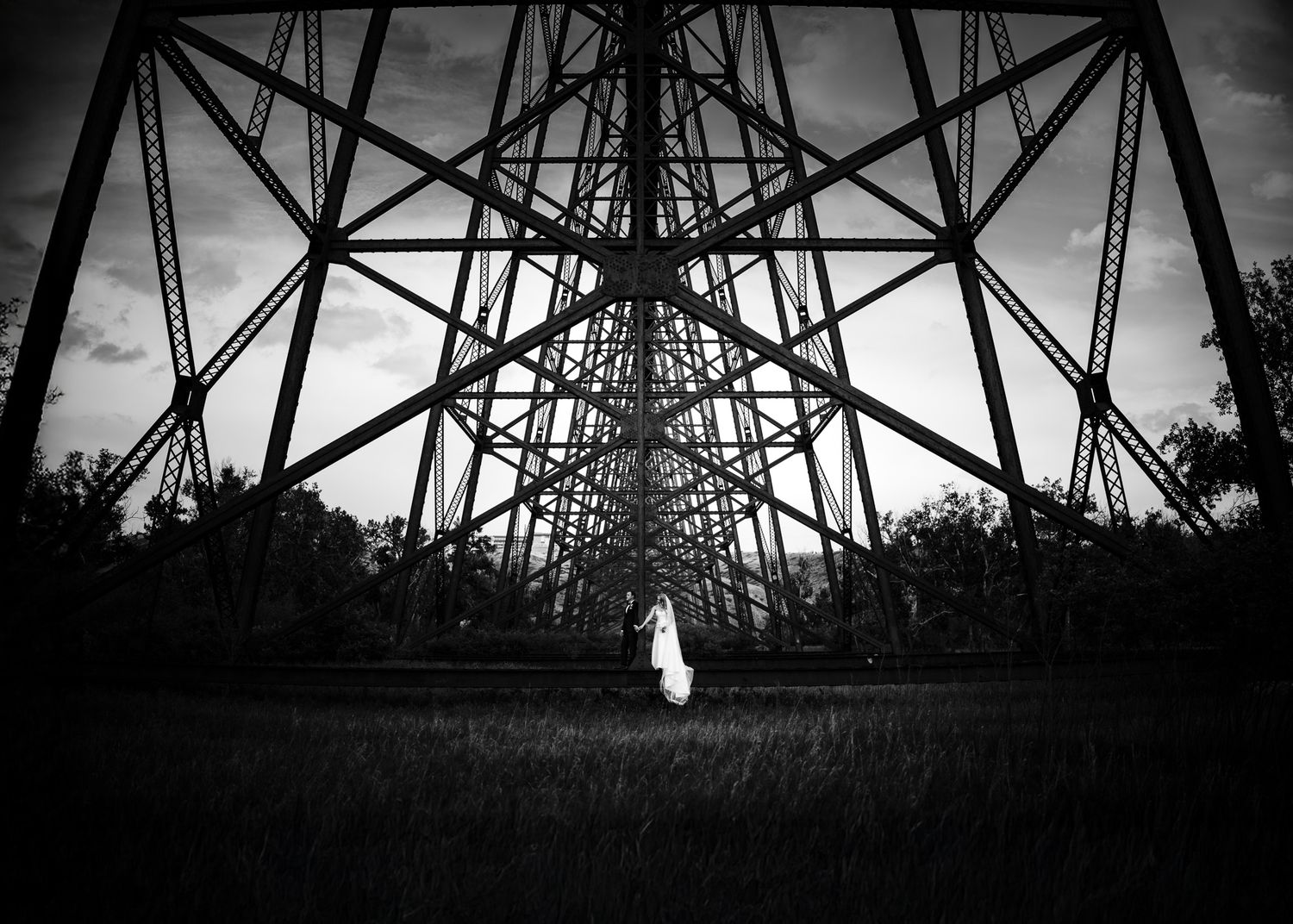 Black and white bride and groom beneath a towering steel trestle bridge, her veil trailing, by a Calgary wedding photographer