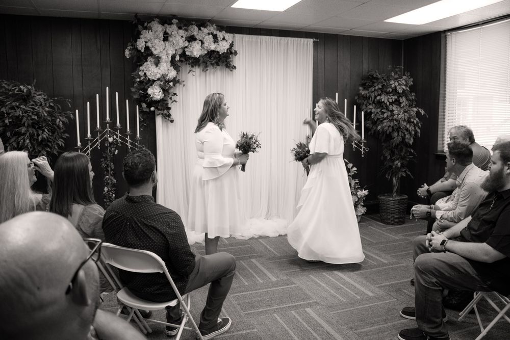 Two brides in white gowns exchange vows at an intimate wedding ceremony with floral arch and seated guests.