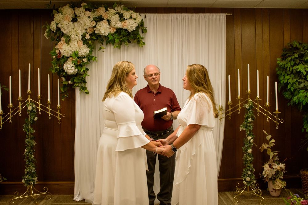 Two brides in white dresses hold hands during wedding ceremony beneath floral arch with officiant.