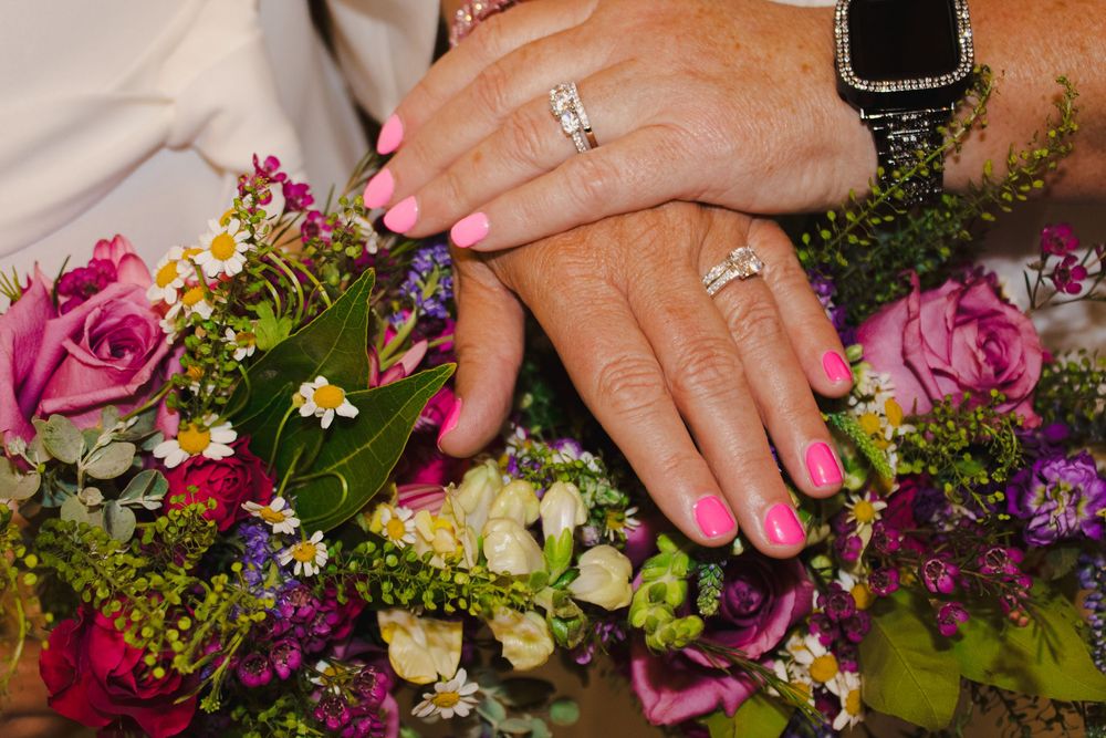 Two brides display wedding rings with pink manicures over a vibrant bouquet of roses and wildflowers.