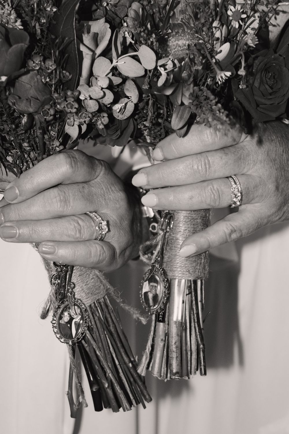 Black and white close-up of two brides holding floral bouquet with charm accessories and wedding rings.