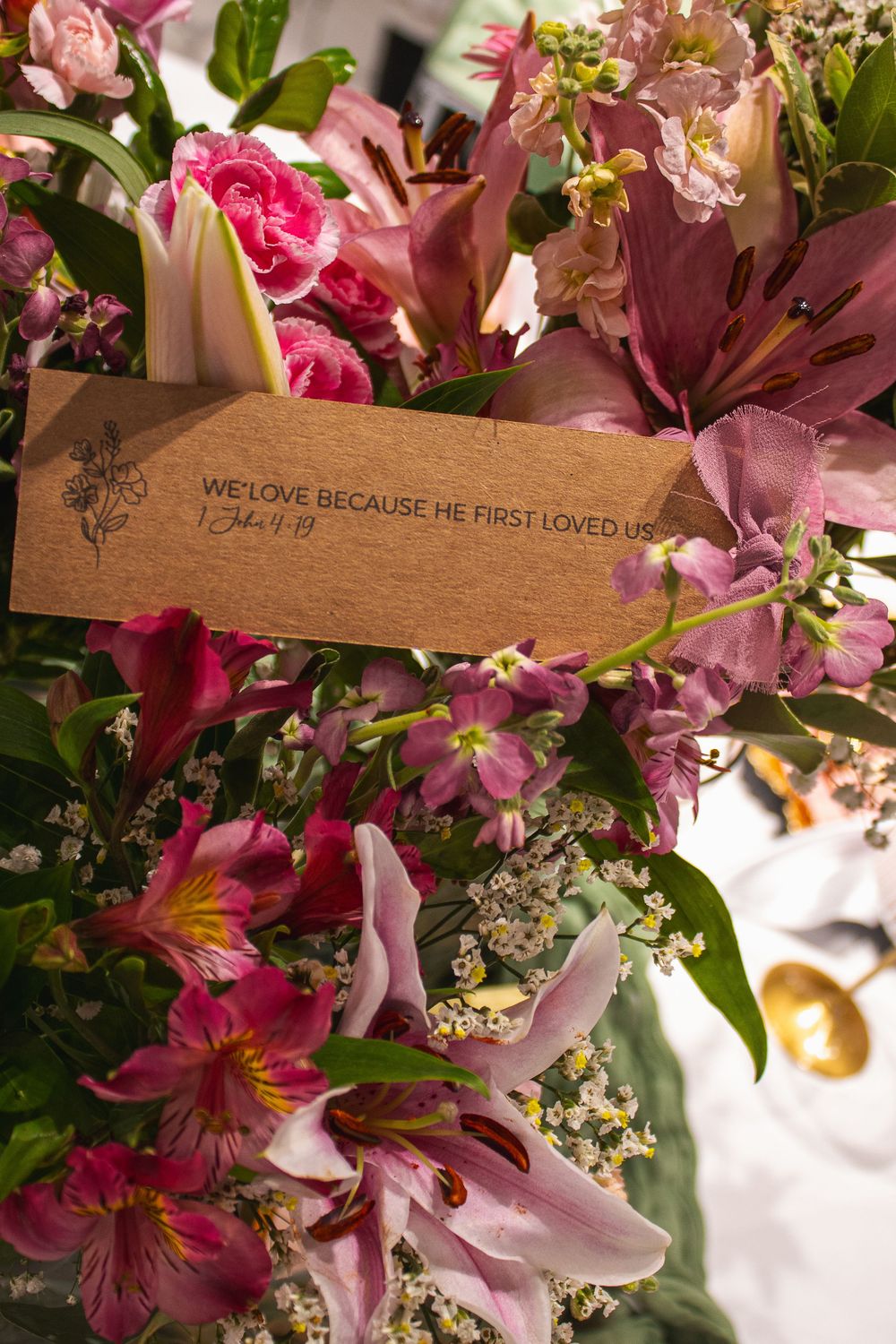 Lush pink floral arrangement with lilies and roses featuring a kraft card with Bible verse 1 John 4:19.