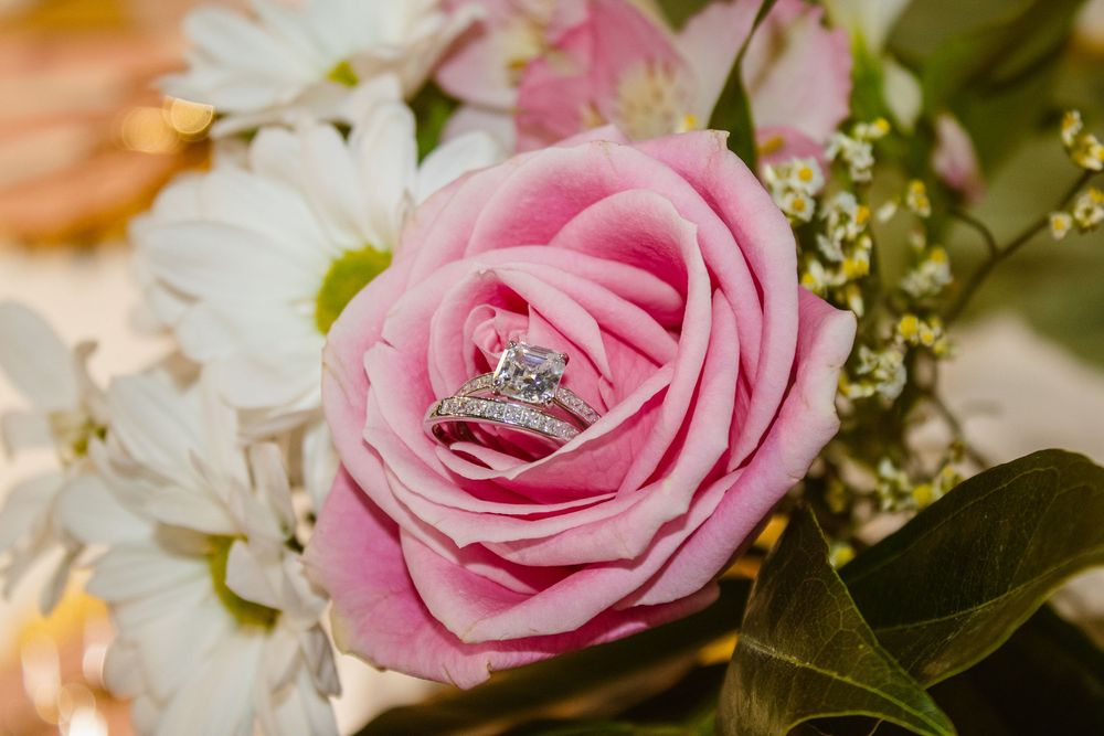 Diamond engagement ring resting inside a pink rose surrounded by white daisies and wildflowers in a bridal bouquet.