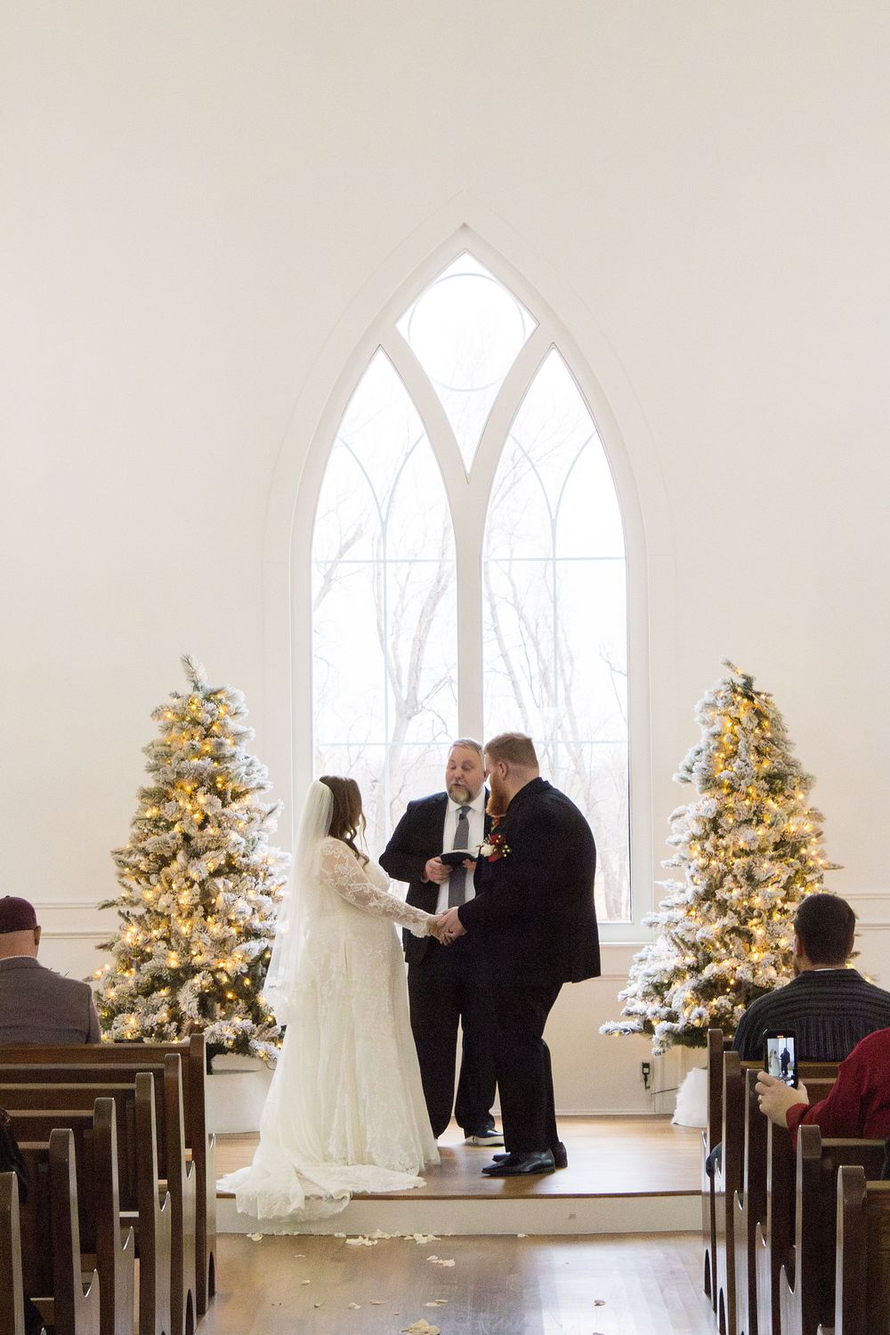 Bride and groom exchange vows at winter chapel wedding altar with decorated Christmas trees and Gothic arch window.
