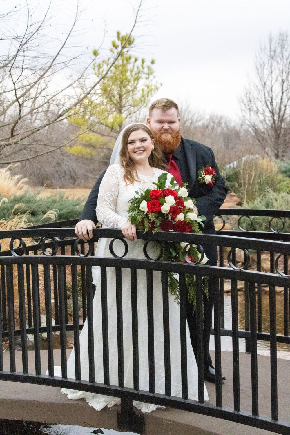 Bride and groom pose on ornate bridge holding red and white bouquet, groom in dark suit with red tie.