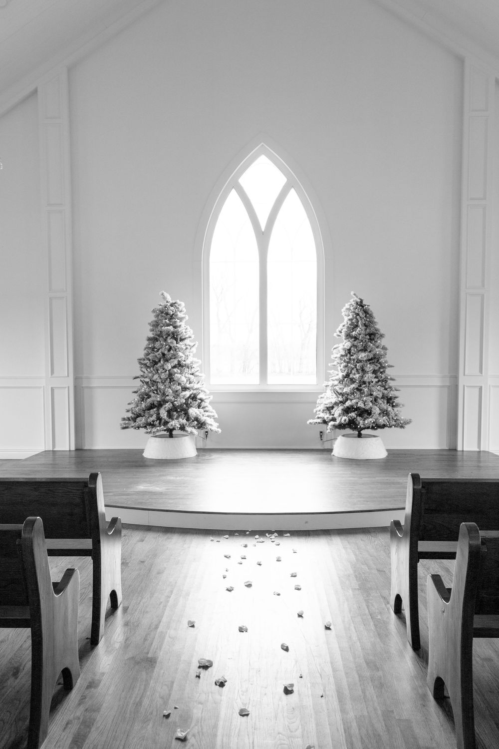 Black and white chapel interior with snow-flocked Christmas trees flanking an altar, wooden pews, and Gothic arched window.