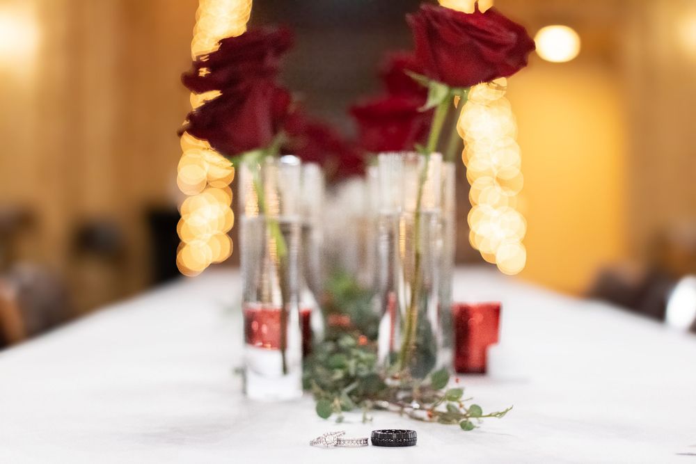 Elegant wedding table centerpiece with red roses in glass vases, greenery, and rings on white linen.
