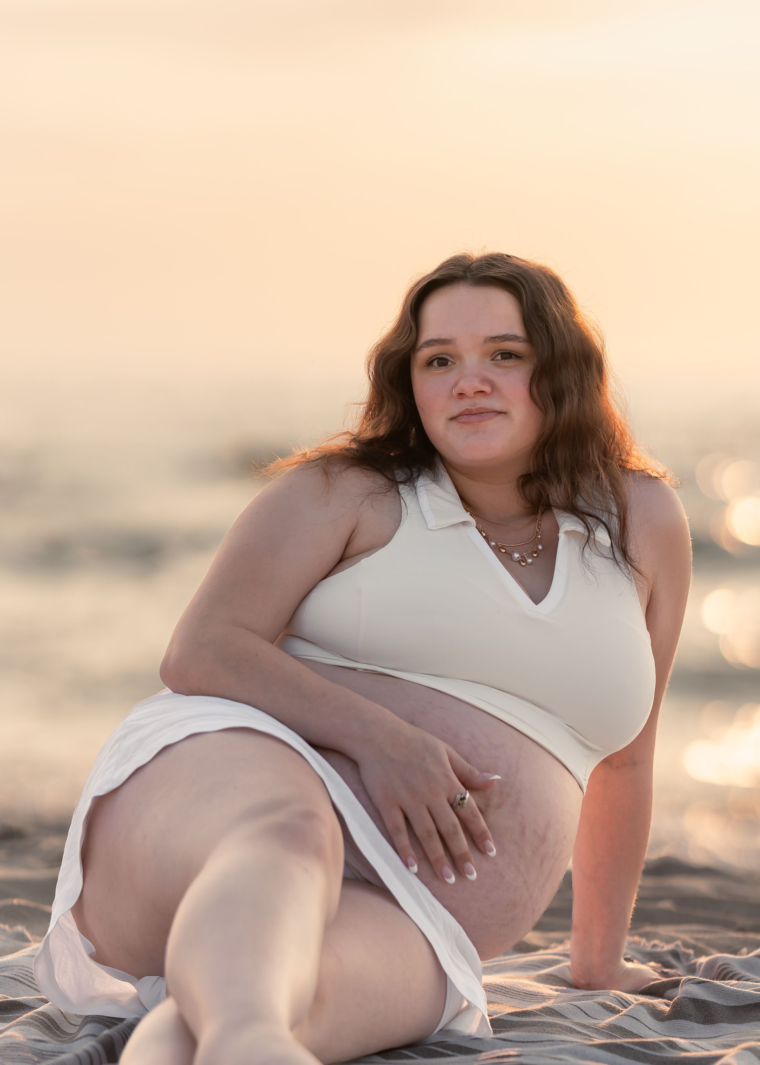 Pregnant woman in white outfit sitting on beach at golden sunset, cradling baby bump with serene expression.