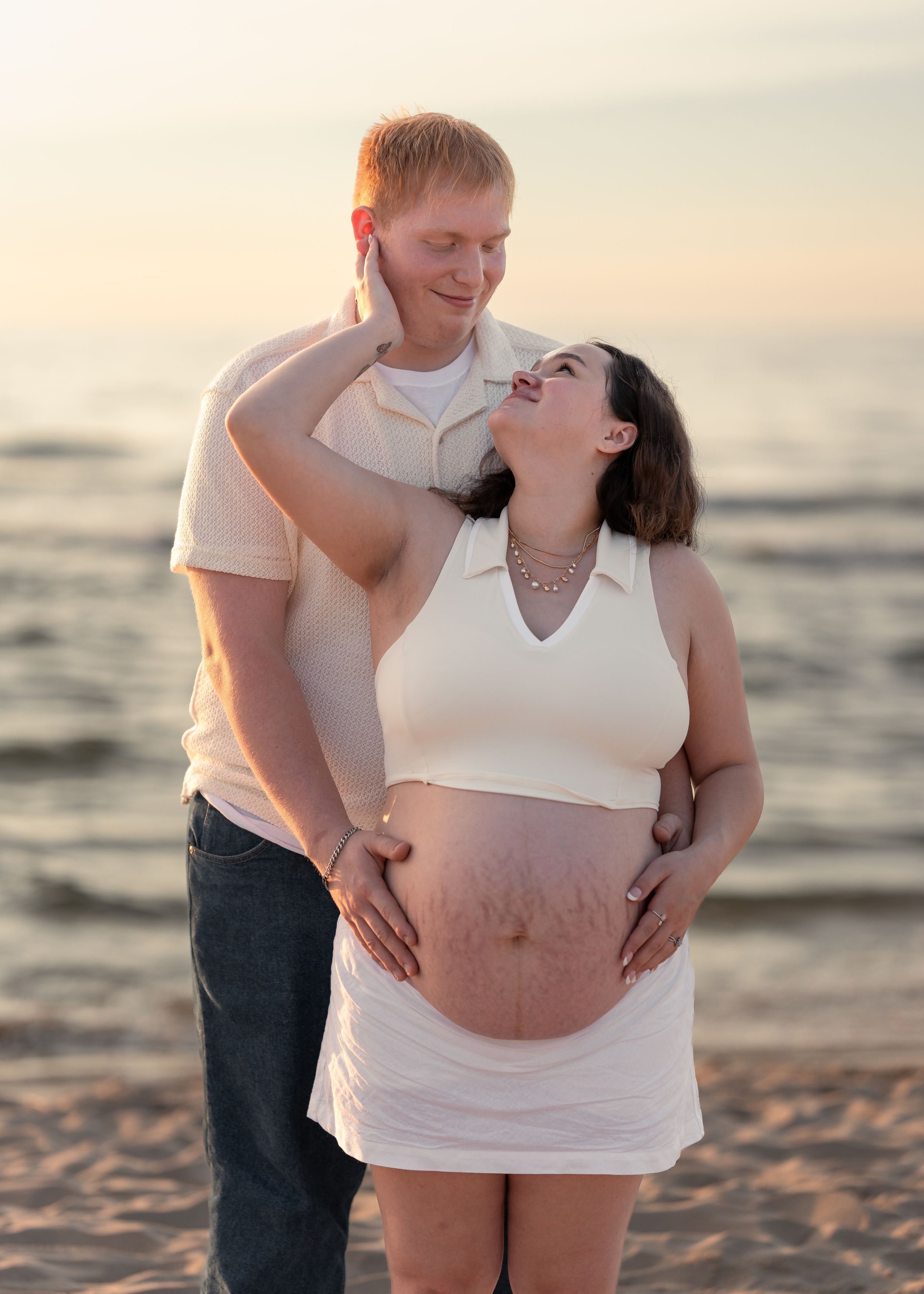 Pregnant couple sharing a tender moment on a beach at sunset, woman cradling her baby bump in a white outfit.