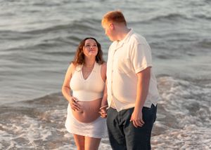 Pregnant couple holding hands and smiling at each other on a beach at sunset, woman cradling baby bump.