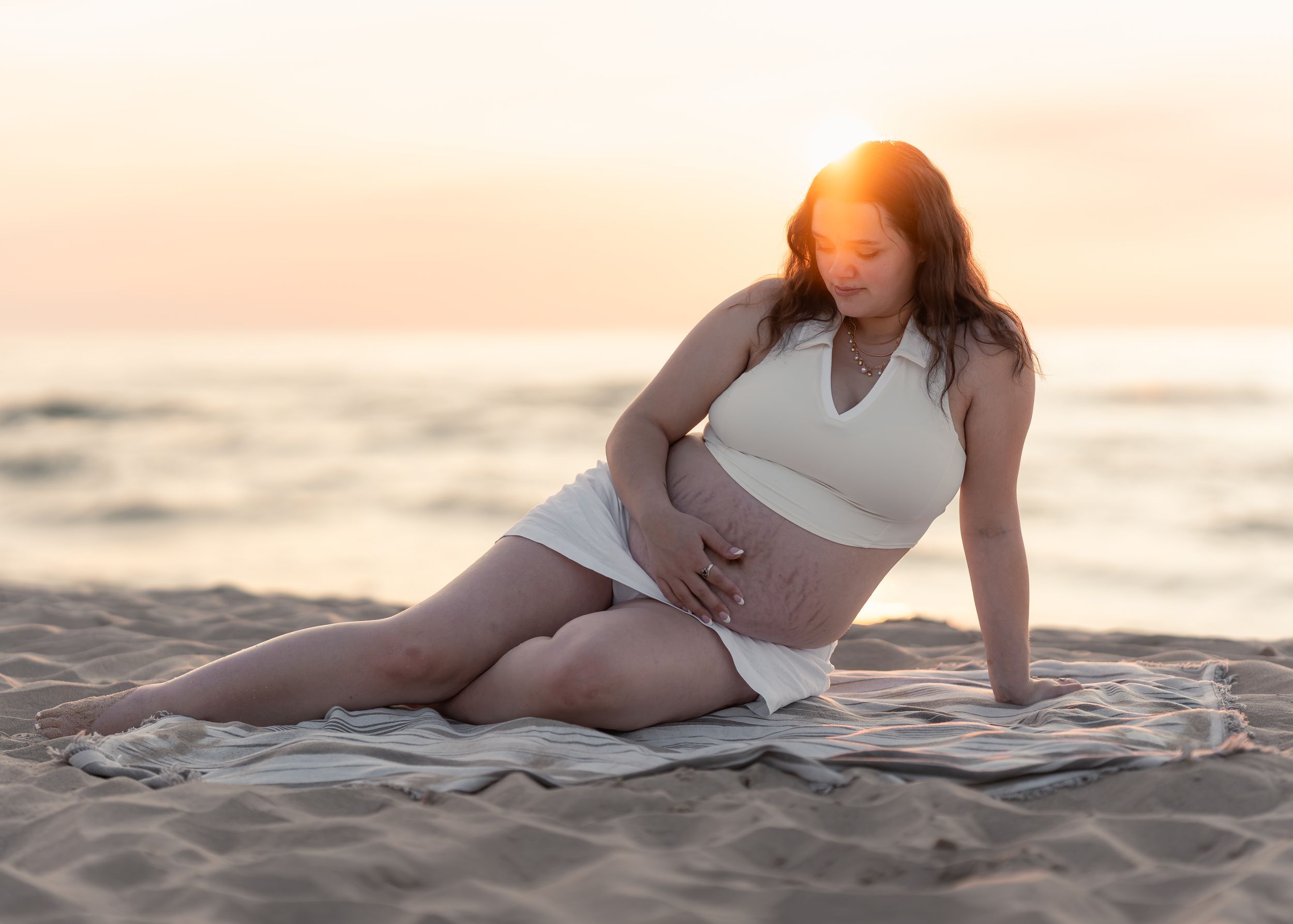 Pregnant woman sitting on beach blanket at sunset, cradling her baby bump in a white crop top and skirt.
