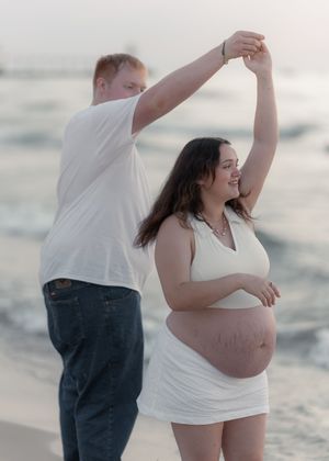 Pregnant couple dancing on beach at sunset, man spinning woman in white dress with baby bump near ocean waves.