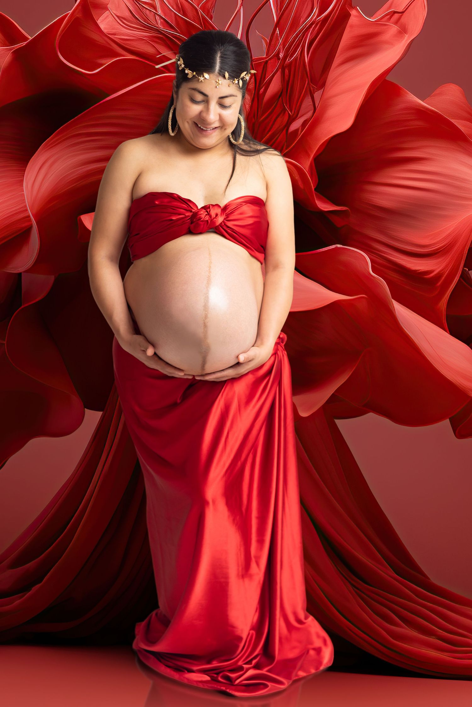 Pregnant woman in red satin gown holding belly, adorned with gold crown, against flowing red fabric backdrop.